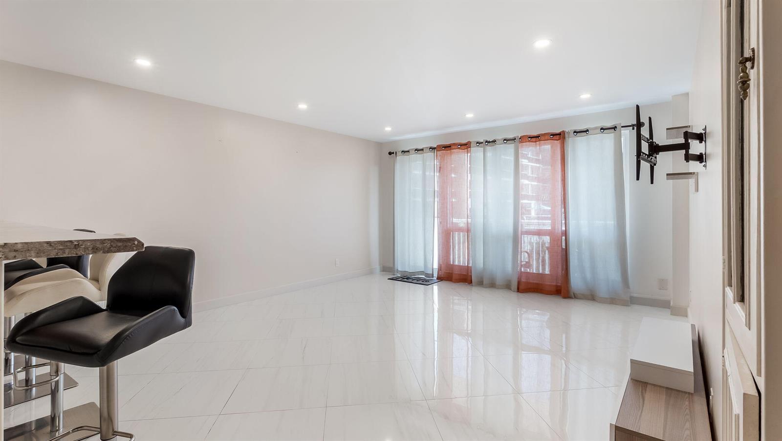 A bright living room features white tile flooring, a wall-mounted TV bracket, and layered curtains — 133 N Pompano Beach Boulevard # 204, Pompano Beach FL 33062, POMPANO BEACH, FL