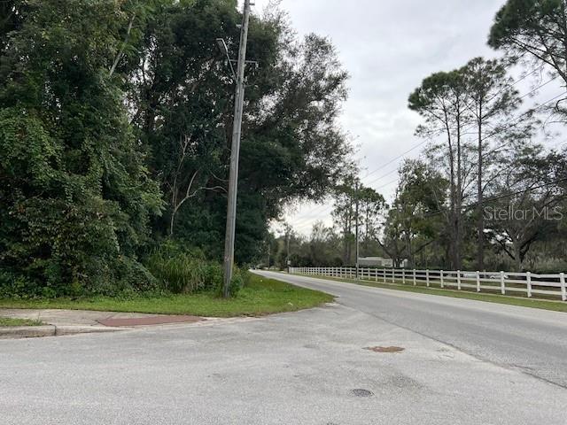 A paved road curves past a grassy corner lot with dense trees and a white fence — 1290 W PLYMOUTH AVENUE, DELAND, FL