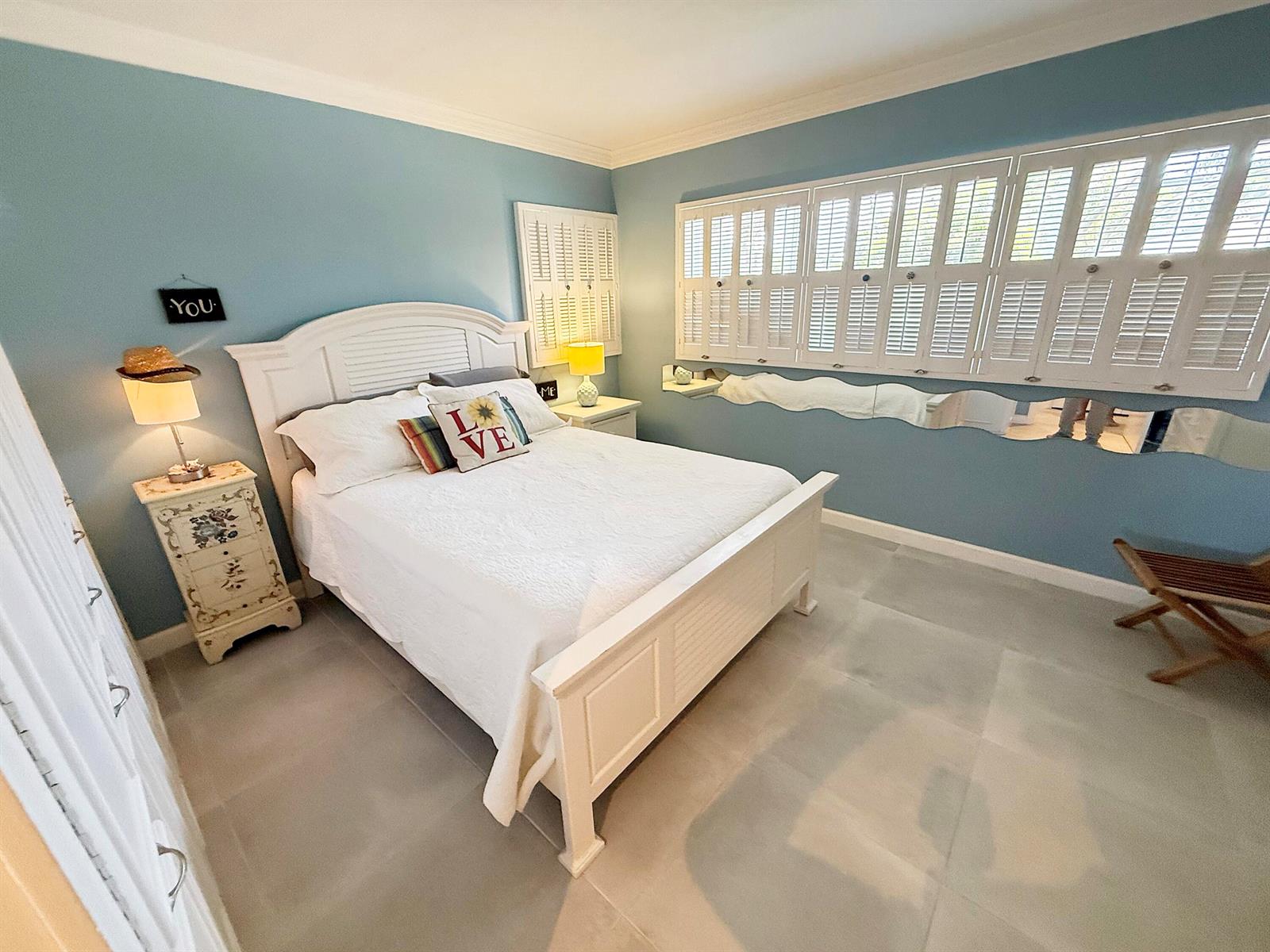 A bedroom features light blue walls, a white bed, tile flooring, and white plantation shutters — 1201 S Riverside Drive # 106, Pompano Beach FL 33062, POMPANO BEACH, FL