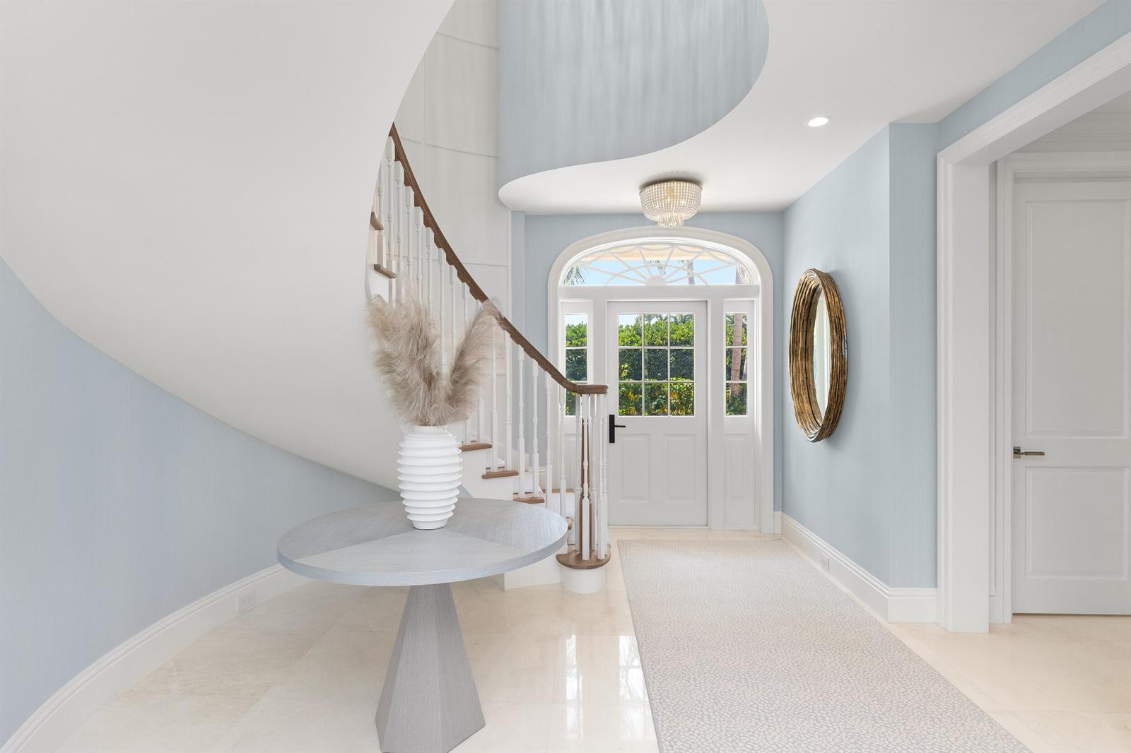 A bright foyer features a curved staircase, light blue walls, and a central pedestal table — 211 Ocean Terrace, Palm Beach FL 33480, PALM BEACH, FL