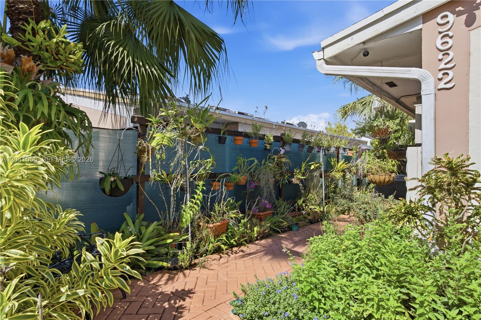 A brick walkway leads along a house exterior lined with numerous potted plants and orchids — 9622 SW 146th Ave, Miami FL 33186, MIAMI, FL