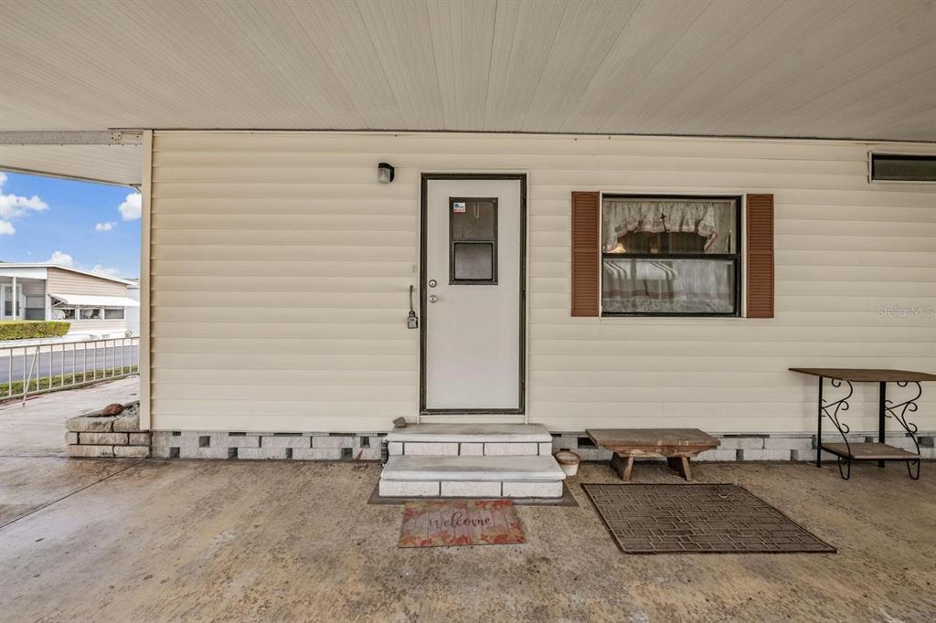 A beige mobile home exterior features a white front door, a window, and a concrete carport — 7047 EL MATADOR STREET, ZEPHYRHILLS, FL