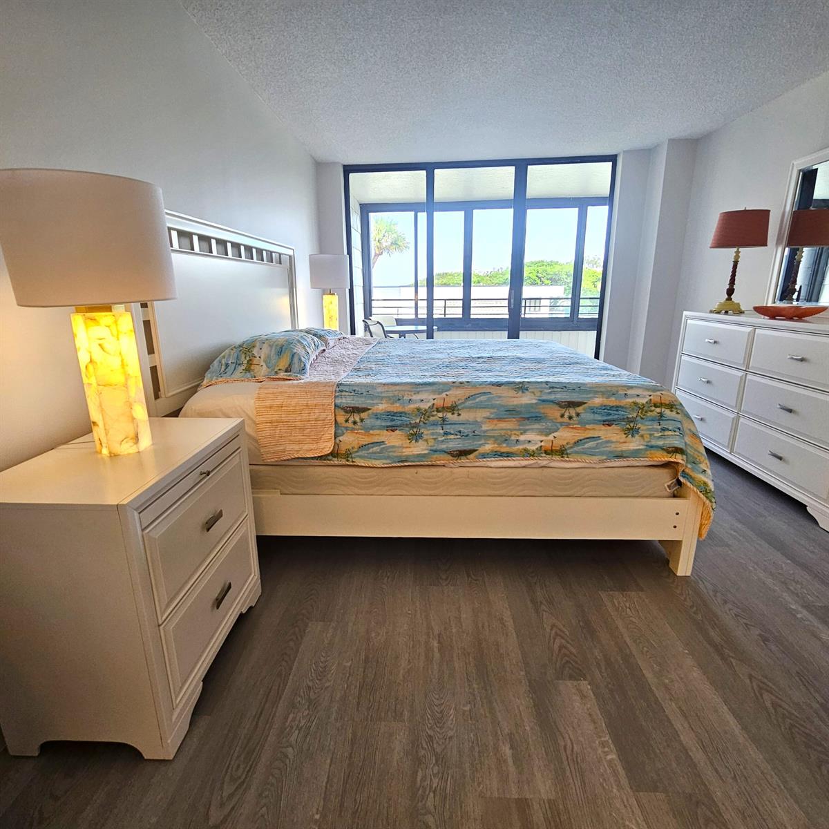 A bedroom features a bed with a tropical quilt, white furniture, and a balcony view — 1357 NE Ocean Boulevard # 215, Stuart FL 34996, STUART, FL