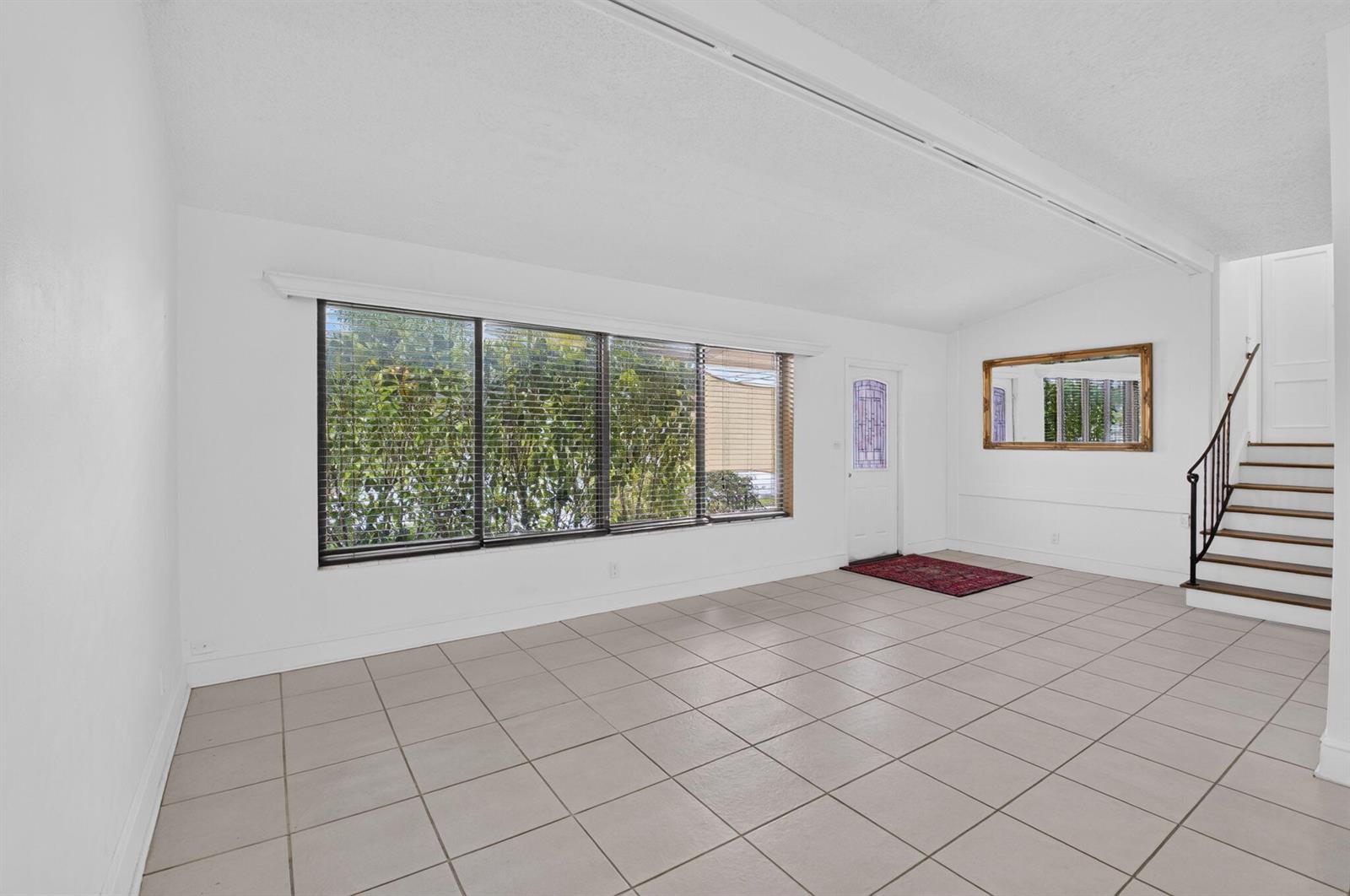 A bright, empty living room features white walls, large windows, tile flooring, and a staircase — 424 NW 12th Avenue, Boca Raton FL 33486, BOCA RATON, FL