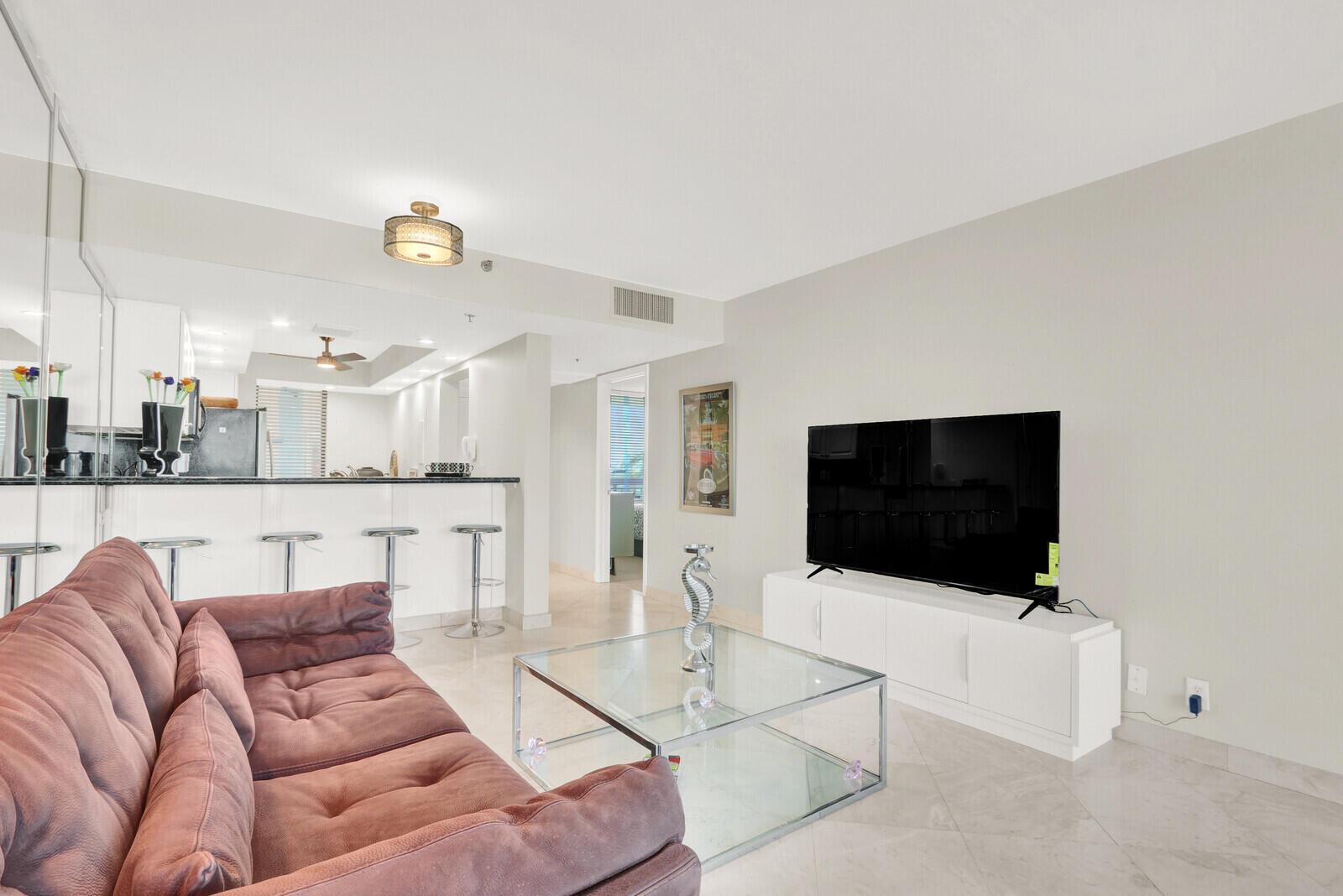 A living room features a mauve sofa, glass coffee table, and a television on a stand — 2687 N Ocean Boulevard # 306, Boca Raton FL 33431, BOCA RATON, FL