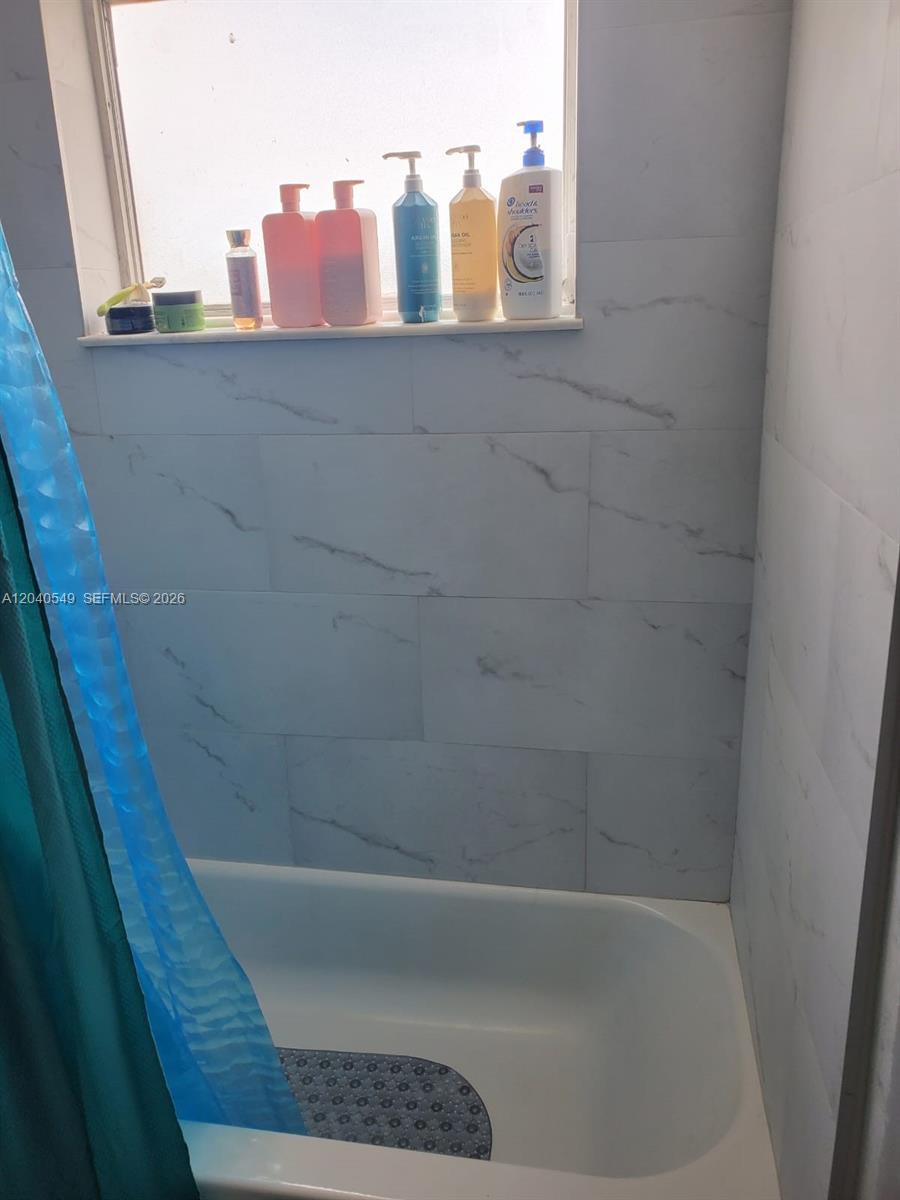 A bathtub with a blue shower curtain and marble-look wall tiles sits below a window — 140 NW 87th Ave # G214, Miami FL 33172, MIAMI, FL