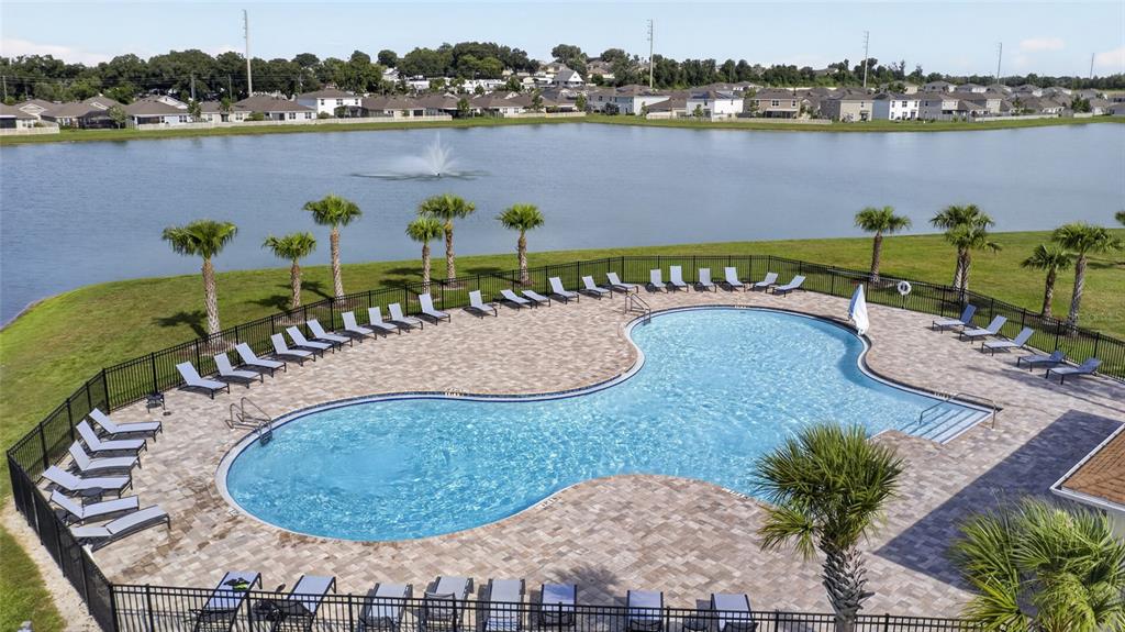 An aerial view shows a swimming pool with lounge chairs on a patio by a lake — 8136 WHEAT STONE DRIVE, ZEPHYRHILLS, FL