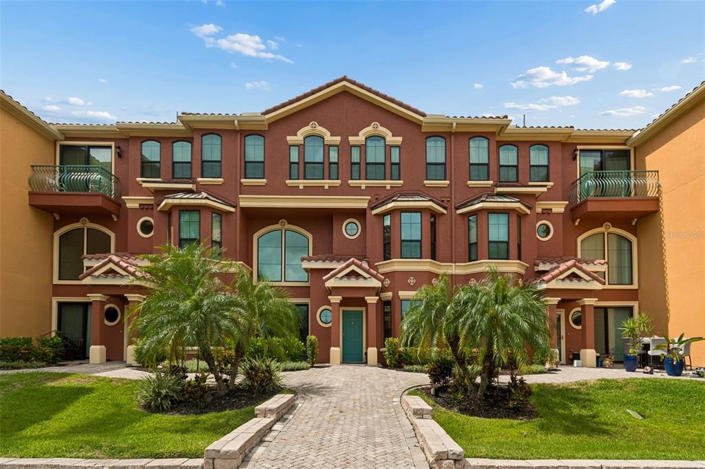 A three-story, reddish-brown apartment building with arched windows, balconies, and a paved walkway — 2717 VIA CIPRIANI UNIT 623A, CLEARWATER, FL