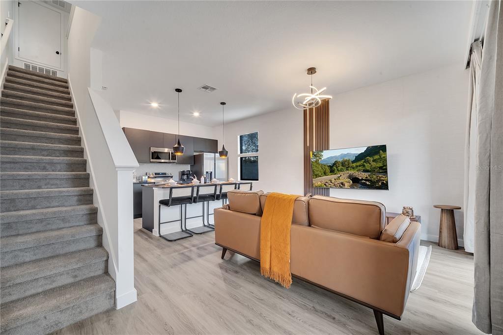 A modern open-concept living room and kitchen featuring a brown sofa, stairs, and dark cabinetry — 8752 GOOD VIBES PLACE, KISSIMMEE, FL