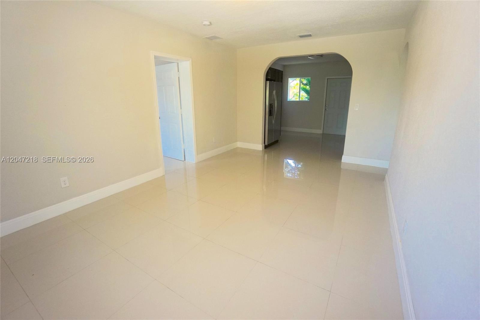 An empty room with light-colored tile flooring leads through an arched doorway into a kitchen — 1497 NE 118th Ter # 2, Miami FL 33161, MIAMI, FL