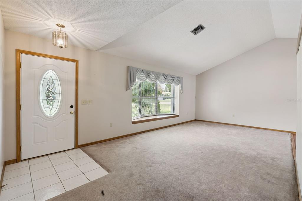 An empty living room features a vaulted ceiling, beige carpeting, a bay window, and tiled entryway — 4160 WOOD DRIVE, MOUNT DORA, FL