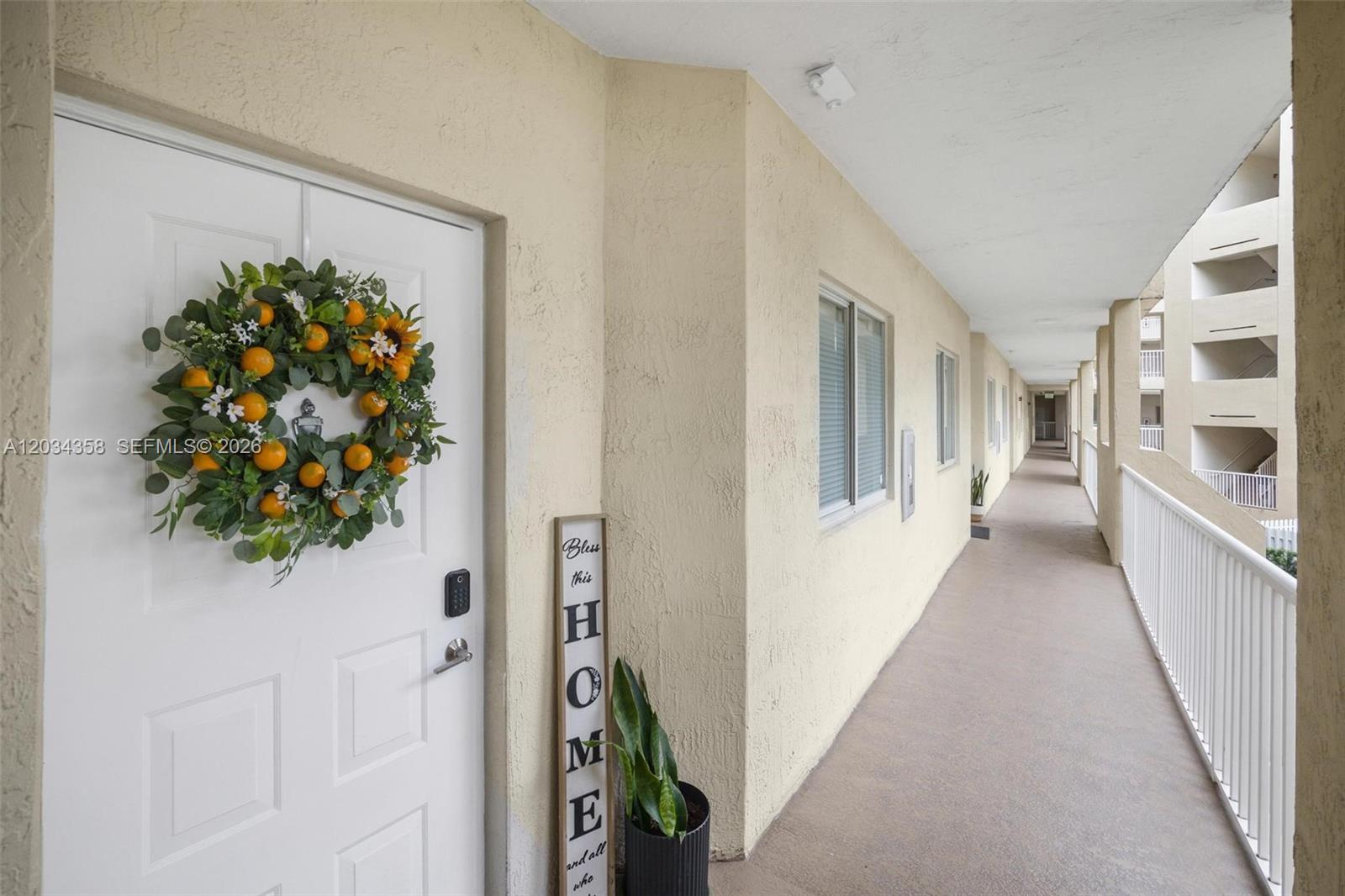 An outdoor apartment hallway features a white door with a wreath and a home sign — 10720 NW 66th St # 202, Doral FL 33178, DORAL, FL