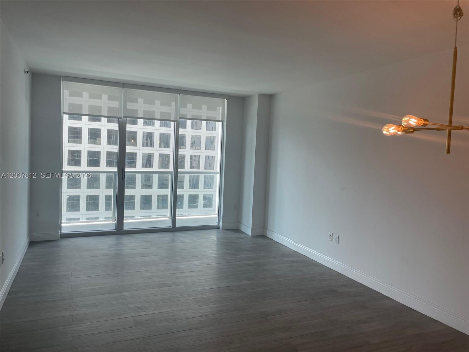 An empty room features gray wood-look flooring, white walls, a modern chandelier, and floor-to-ceiling windows — 500 Brickell Av # 1906, Miami FL 33131, MIAMI, FL