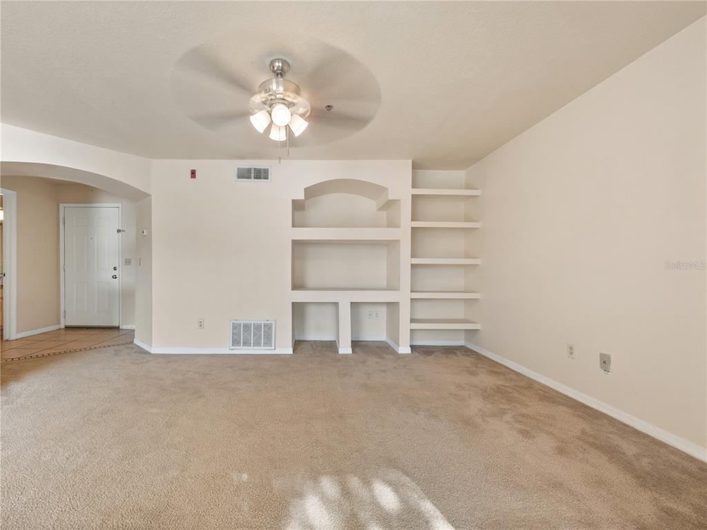A carpeted living room features a ceiling fan, built-in shelving, and an arched entryway — 7210 WESTPOINTE BOULEVARD UNIT 1325, ORLANDO, FL
