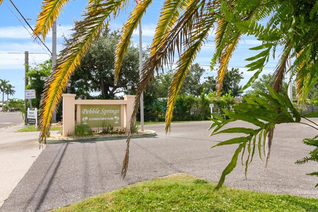The entrance sign for Pebble Springs Condominium stands near a road under palm tree fronds — 6108 7TH AVENUE W UNIT 6108, BRADENTON, FL