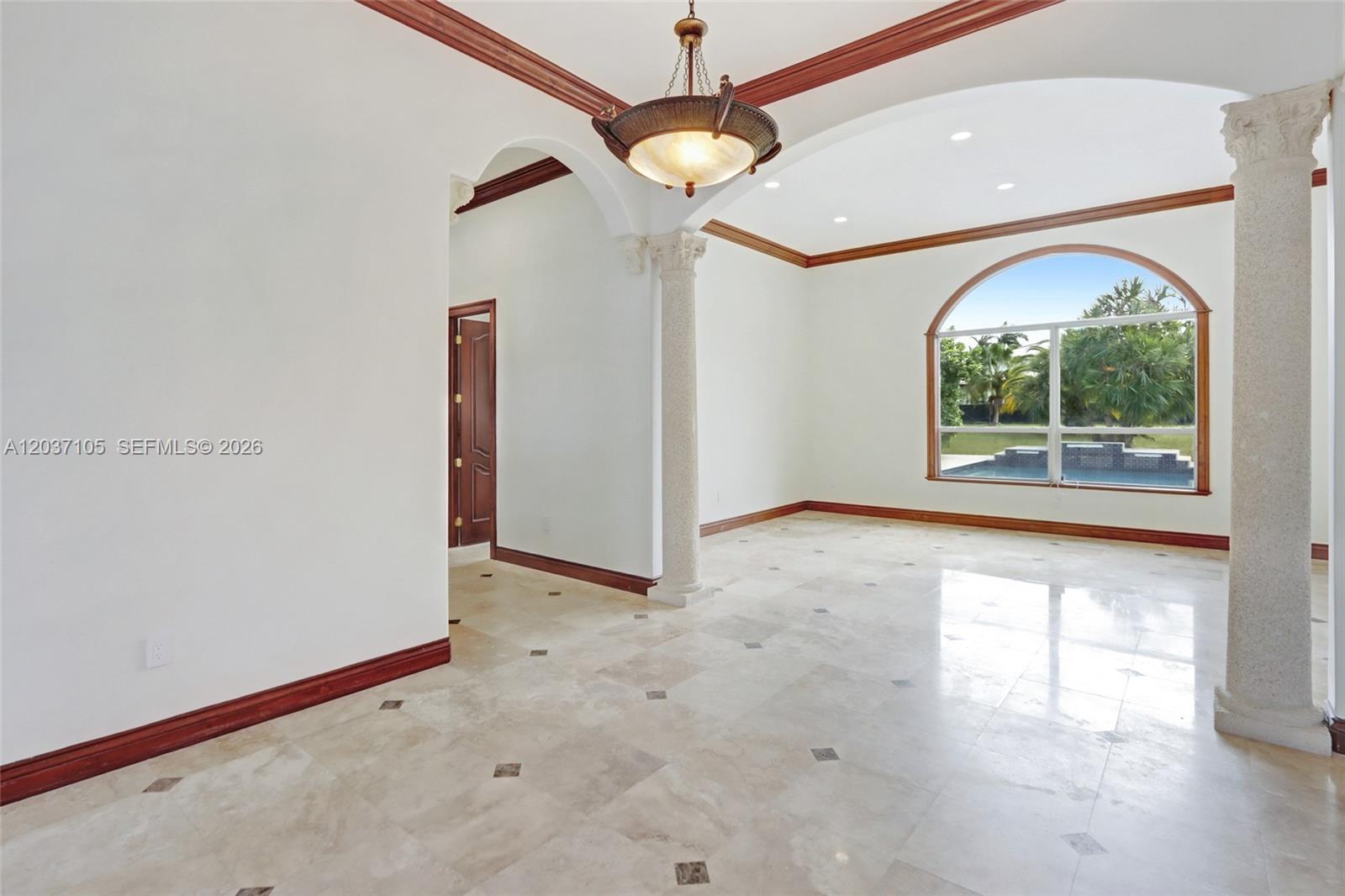 An empty room features stone tile floors, decorative columns, crown molding, and a large arched window — 8480 SW 98th St, Miami FL 33156, MIAMI, FL