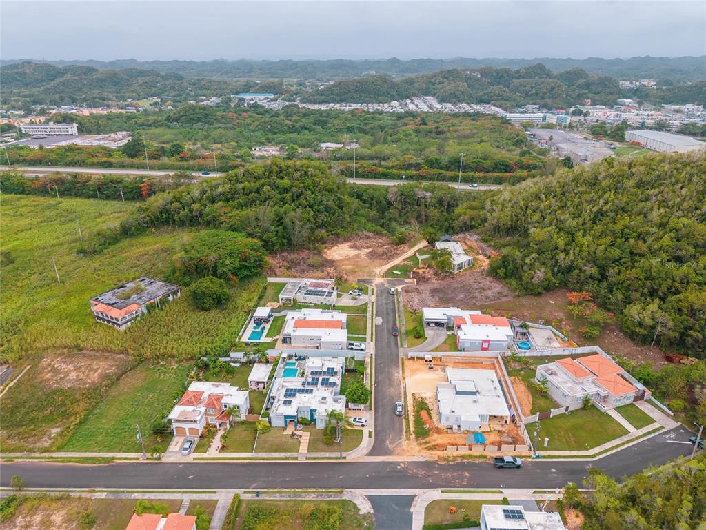 An aerial view shows a suburban neighborhood with several houses, a paved road, and hills — URB. REPARTO ROSELLO, MANATI, PR