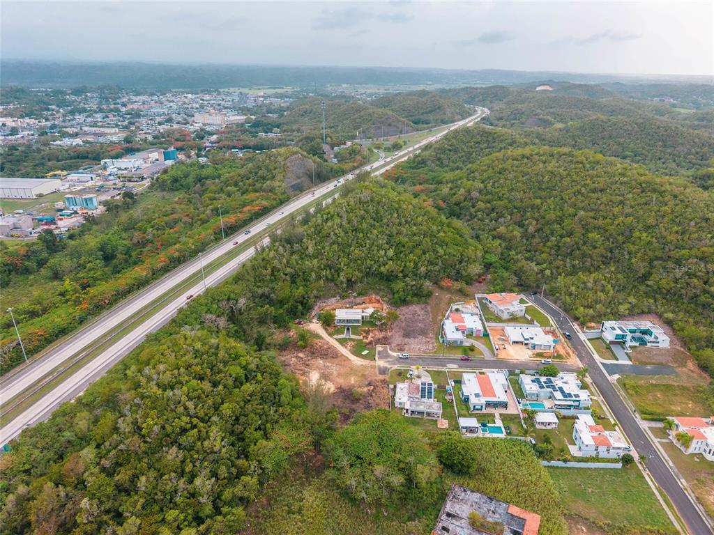 An aerial view shows a residential neighborhood nestled in a hilly, forested landscape beside a highway — URB. REPARTO ROSELLO, MANATI, PR