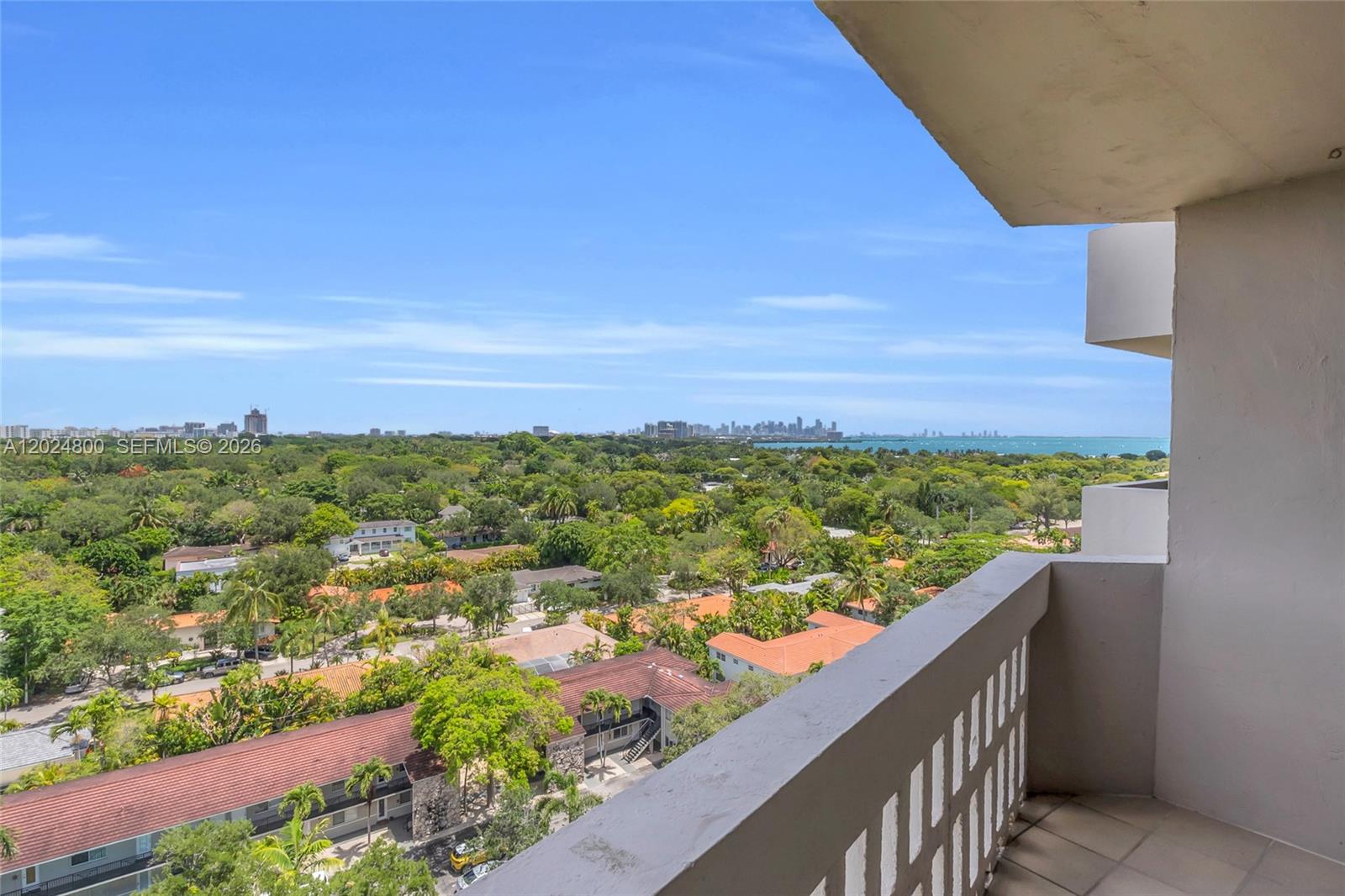 A high-angle view from a balcony overlooks a lush neighborhood and a distant city skyline — 90 Edgewater Dr # 1201, Coral Gables FL 33133, CORAL GABLES, FL