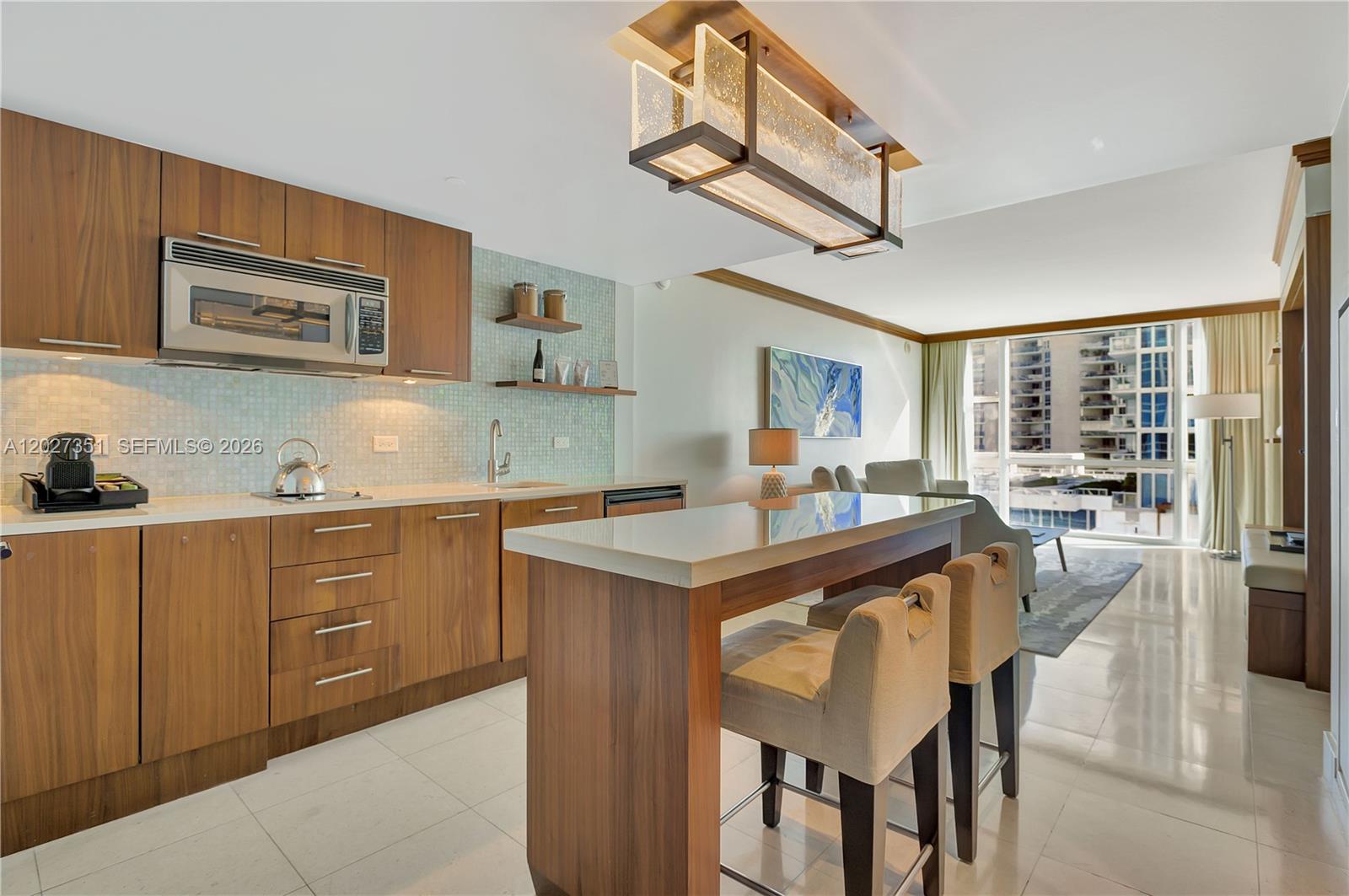 A modern kitchen with wood cabinets and a breakfast bar opens into a living area — 6801 Collins Ave # 806, Miami Beach FL 33141, MIAMI BEACH, FL