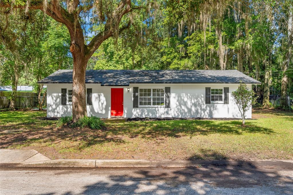 A single-story white house with a bright red front door sits under large oak trees — 2100 SE 50TH STREET, GAINESVILLE, FL
