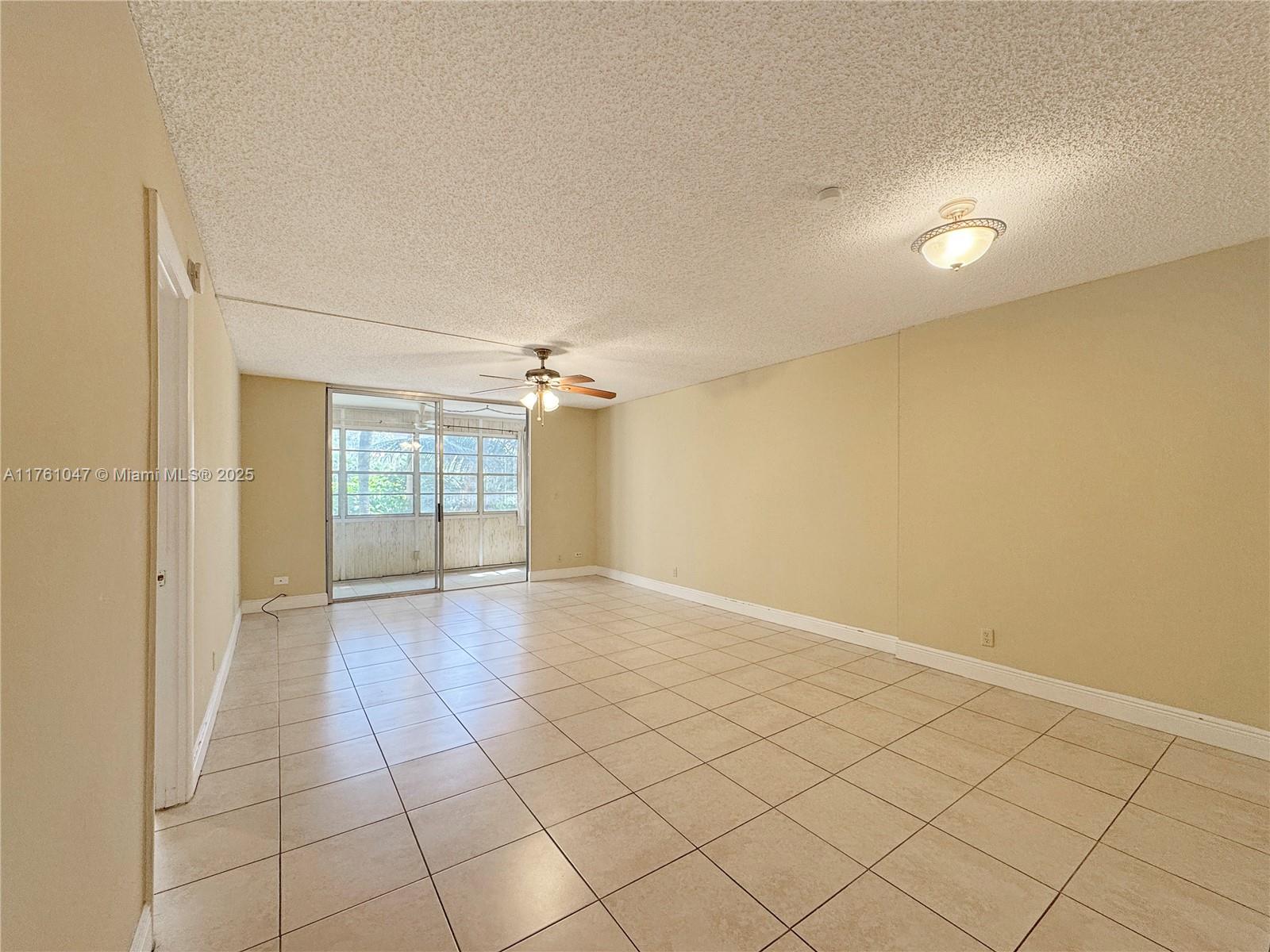 An empty living room features beige tile flooring, a popcorn ceiling, and a sliding glass door — 410 NW 68th Ave # 207, Plantation FL 33317, PLANTATION, FL