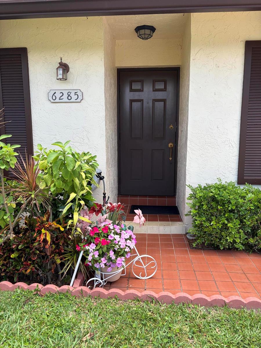 A brown front door is centered on a tiled porch with a decorative bicycle planter — 6285 Tall Cypress Circle, FL 33463, FL