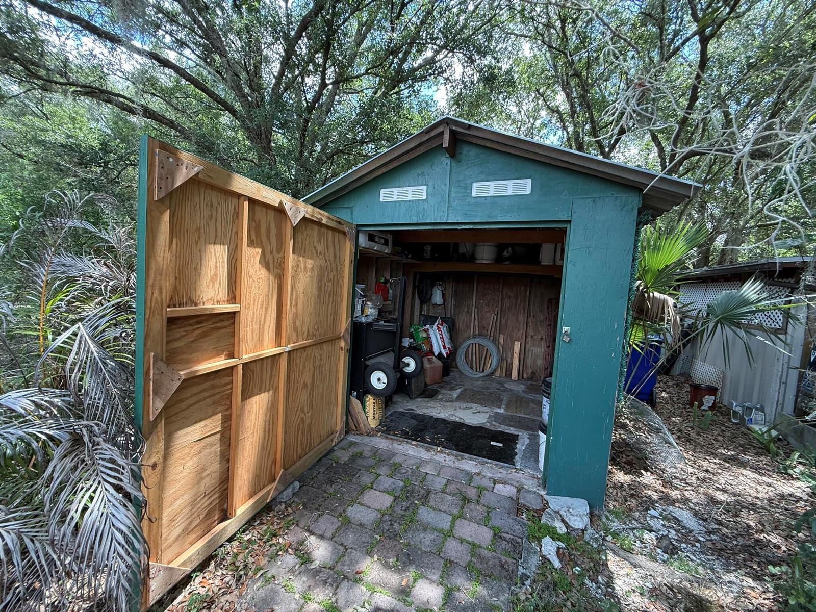 A green wooden shed with an open door sits on a brick patio among trees — 18561 NW 306th Street, Okeechobee FL 34972, OKEECHOBEE, FL
