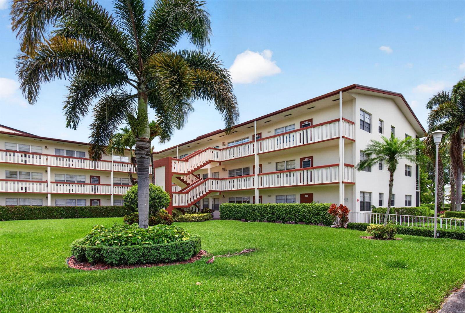 A three-story apartment building with exterior walkways and stairs sits behind a manicured lawn — 374 Brighton I # 374, Boca Raton FL 33434, BOCA RATON, FL