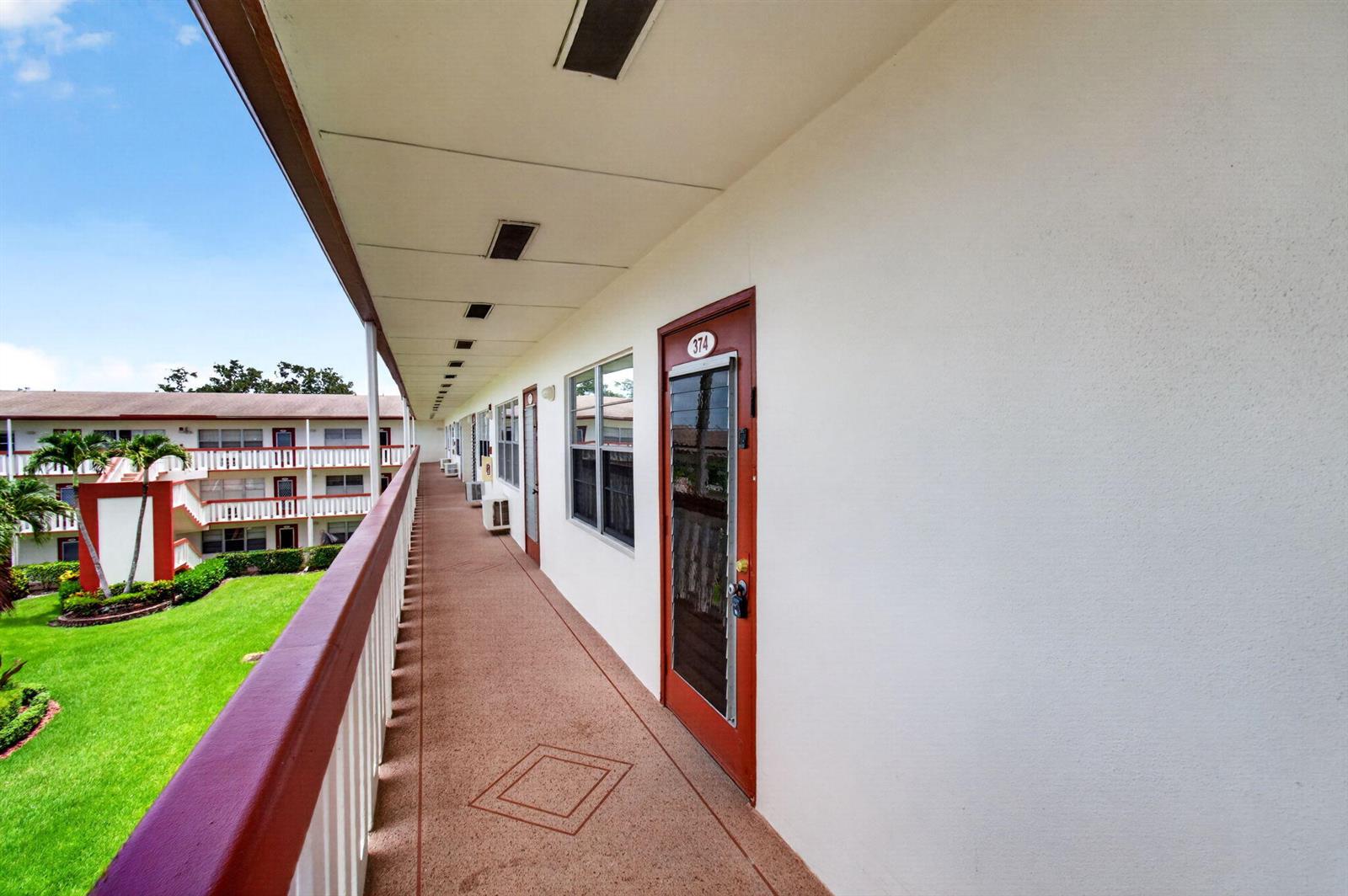 An exterior walkway with red doors and railings overlooks a grassy courtyard — 374 Brighton I # 374, Boca Raton FL 33434, BOCA RATON, FL
