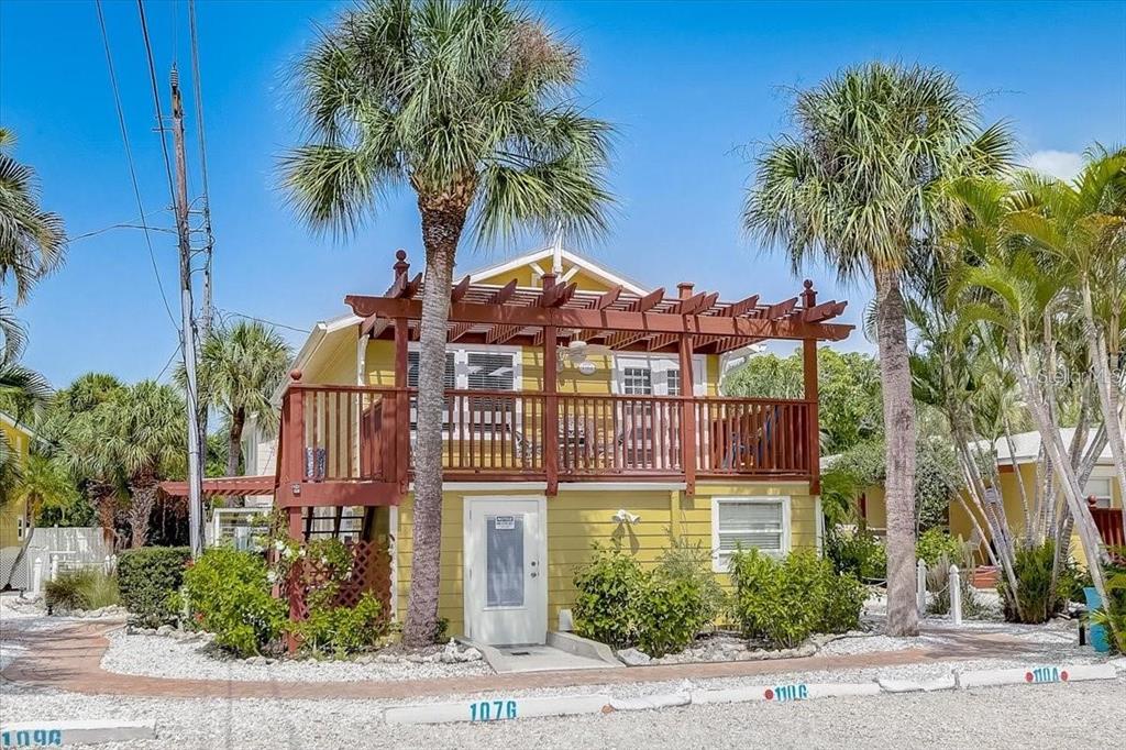 A yellow two-story house features a wooden pergola over a second-story balcony and palm trees — 110 OAK AVENUE UNIT A, ANNA MARIA, FL