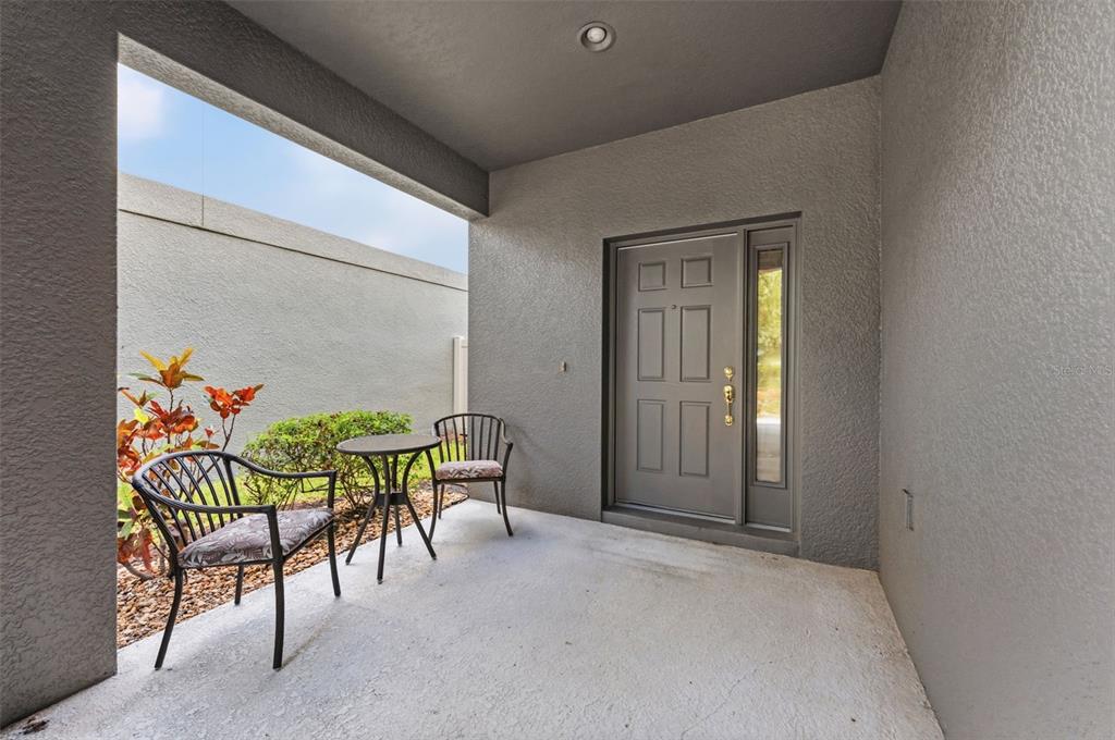 A covered concrete patio features a gray front door, two chairs, and a small table — 10257 NEWMINSTER LOOP, RUSKIN, FL