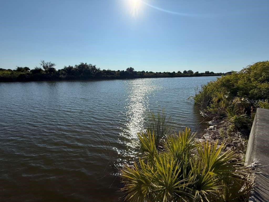A view of a sunlit lake with a rocky shoreline and native vegetation — 13421 GREENCASTLE AVENUE, PORT CHARLOTTE, FL