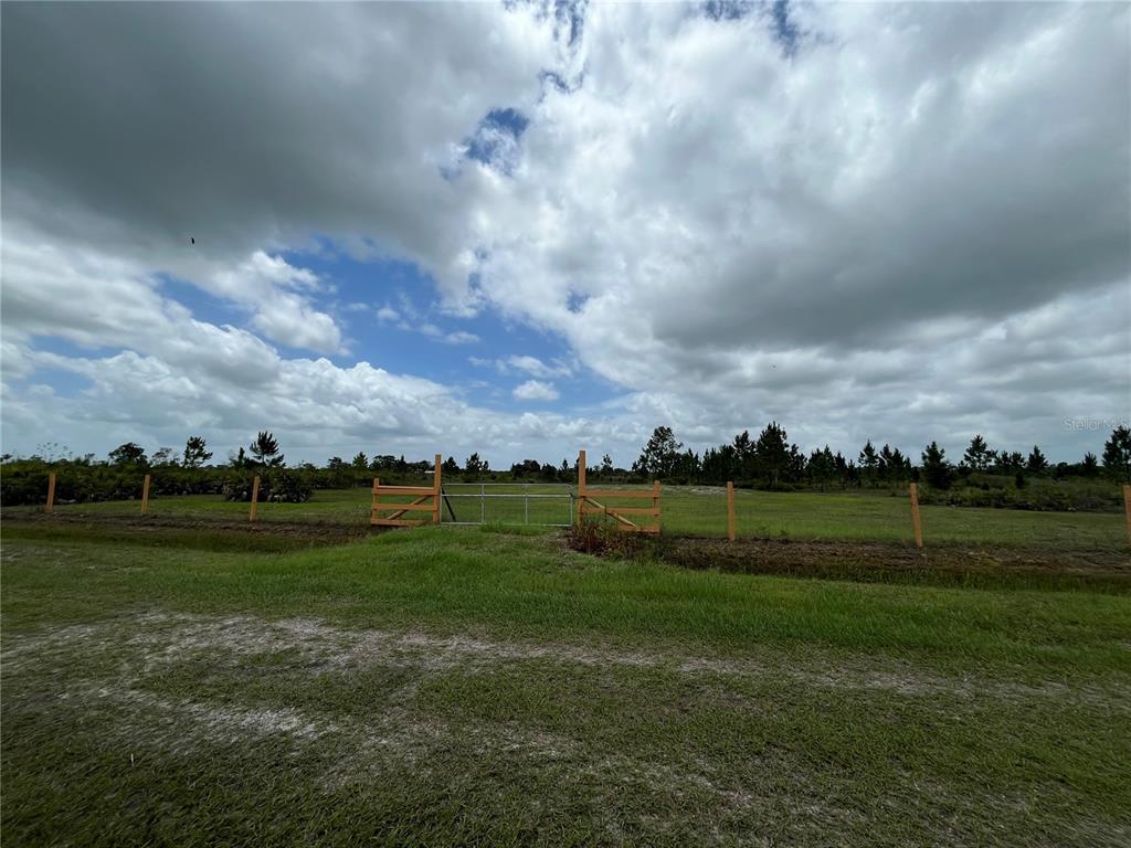 A metal gate sits in a wooden fence line bordering a grassy, open field — 19092 NW 250TH STREET, OKEECHOBEE, FL