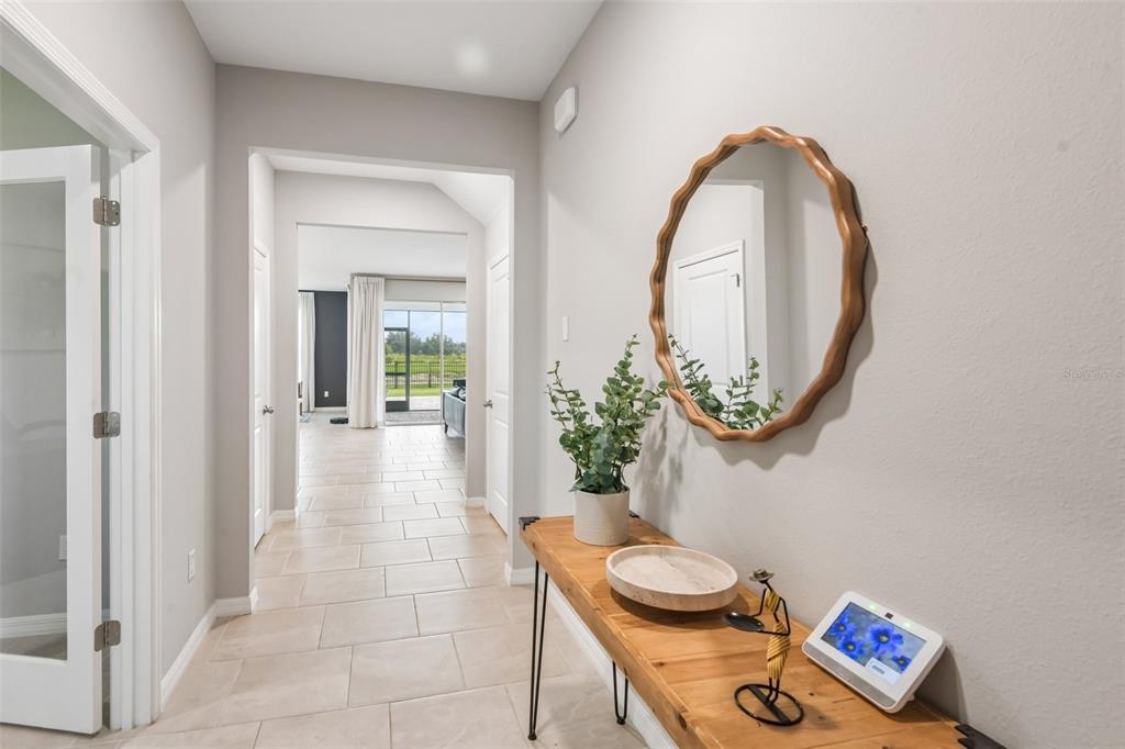 A hallway features light tile flooring, a wooden console table with decor, and a round mirror — 6076 WANDERING WILLOW DRIVE, WESLEY CHAPEL, FL