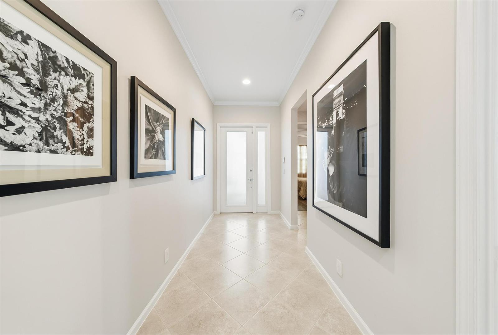 A hallway with tiled floors, neutral walls, and framed black-and-white artwork leads to a door — 2737 Clipper Circle, West Palm Beach FL 33411, WEST PALM BEACH, FL