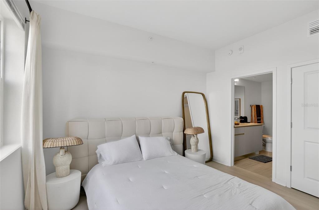 A bedroom features a tufted headboard, two bedside lamps, a floor mirror, and an attached bathroom — 5579 MILLENIA PARK BOULEVARD UNIT 401, ORLANDO, FL