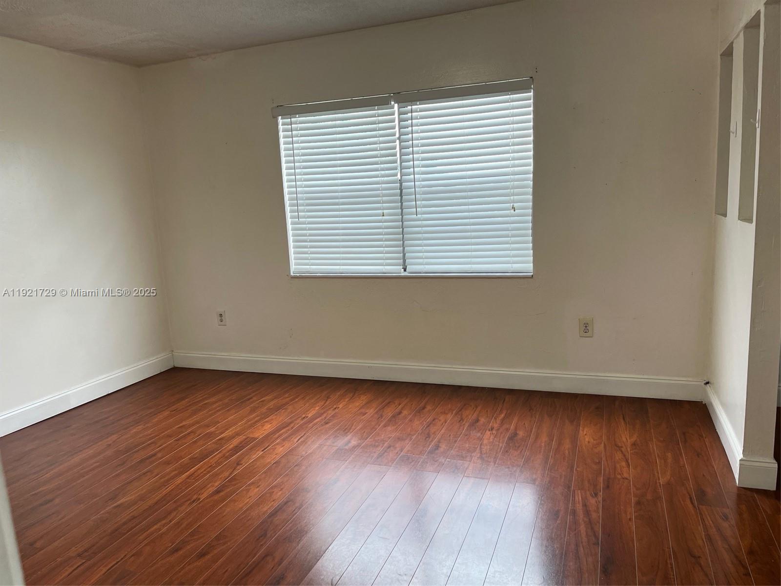 An empty room features white walls, a window with blinds, and dark wood-look flooring — 13500 NE 3rd Ct # 402, North Miami FL 33161, NORTH MIAMI, FL