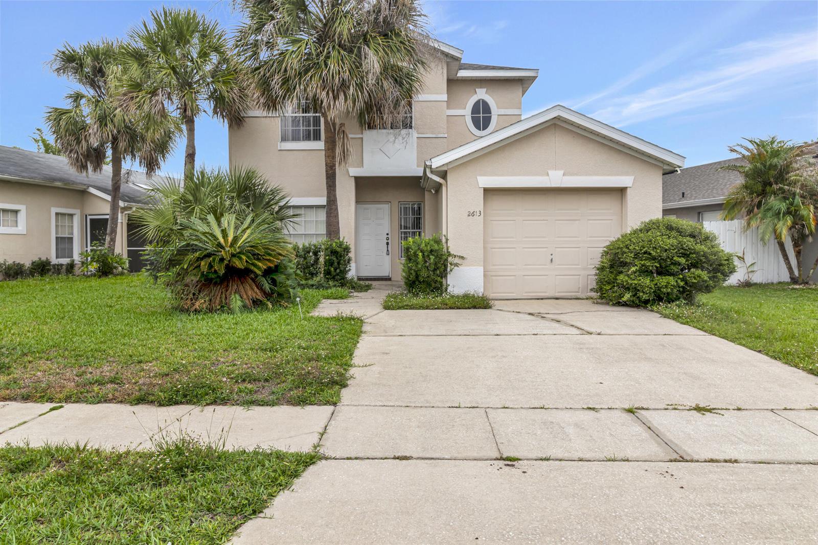 A two-story stucco house features a single-car garage, a concrete driveway, and palm trees — 2613 Spring Hill Drive, Kissimmee FL 34743, KISSIMMEE, FL