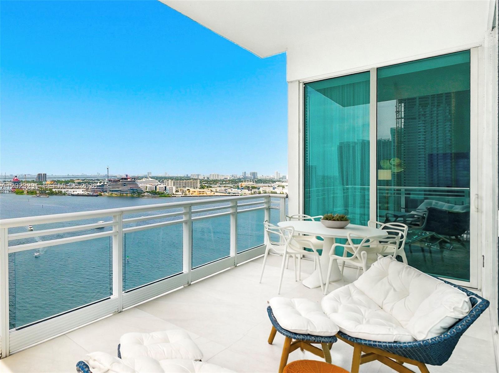 A high-rise balcony features white patio furniture overlooking a bay with a distant cityscape — 900 Brickell Key Blvd # 1801, Miami FL 33131, MIAMI, FL