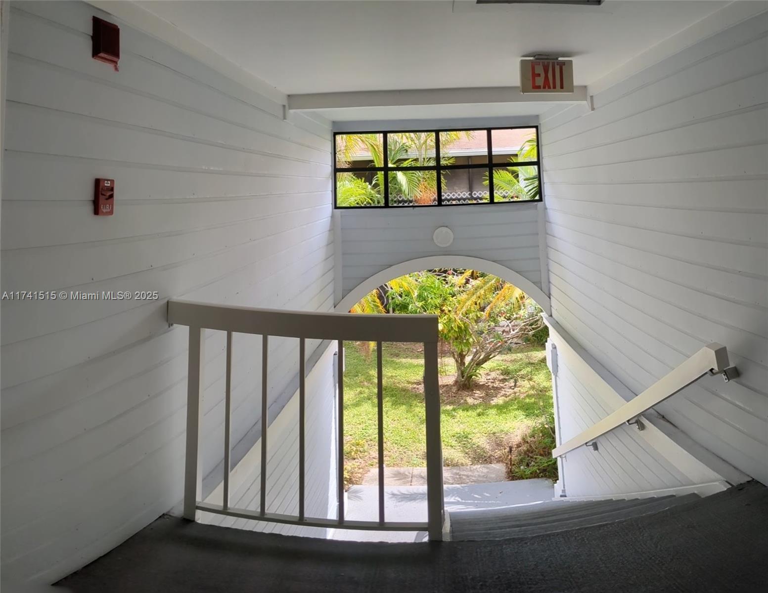 A white-walled stairwell features a wooden railing, an exit sign, and an arched view outdoors — 917 NE 199th St # 206, Miami FL 33179, MIAMI, FL