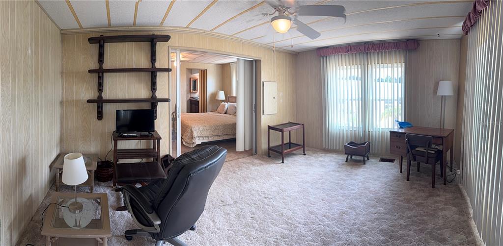 A living room with wood-paneled walls, beige carpet, a ceiling fan, and a view into a bedroom — 3301 ALT 19 UNIT 167, DUNEDIN, FL