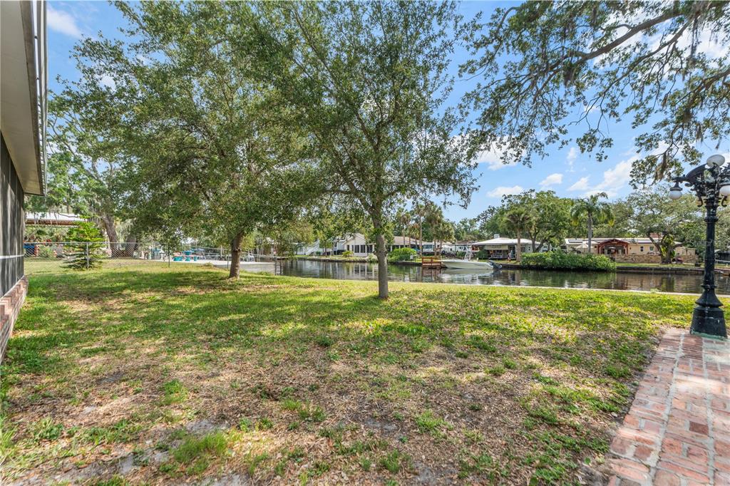 A grassy backyard features mature trees and a brick walkway overlooking a canal with boats — 5822 RIVER ROAD, NEW PORT RICHEY, FL
