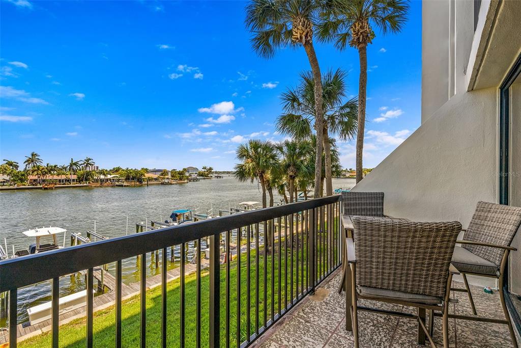 A balcony with wicker furniture overlooks a waterway with boat docks and palm trees — 9495 BLIND PASS ROAD UNIT 202, ST PETE BEACH, FL