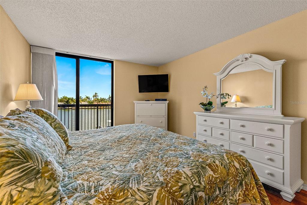 A bedroom features a bed with a tropical print quilt, white furniture, and a balcony view — 9495 BLIND PASS ROAD UNIT 202, ST PETE BEACH, FL