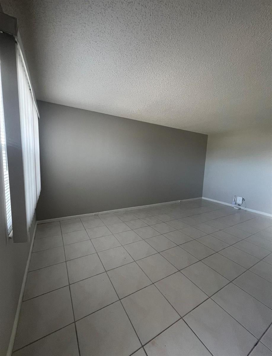 An empty room features light-colored tile flooring, a gray accent wall, and vertical window blinds — 3050 Wolverton C # 3050, Boca Raton FL 33434, BOCA RATON, FL