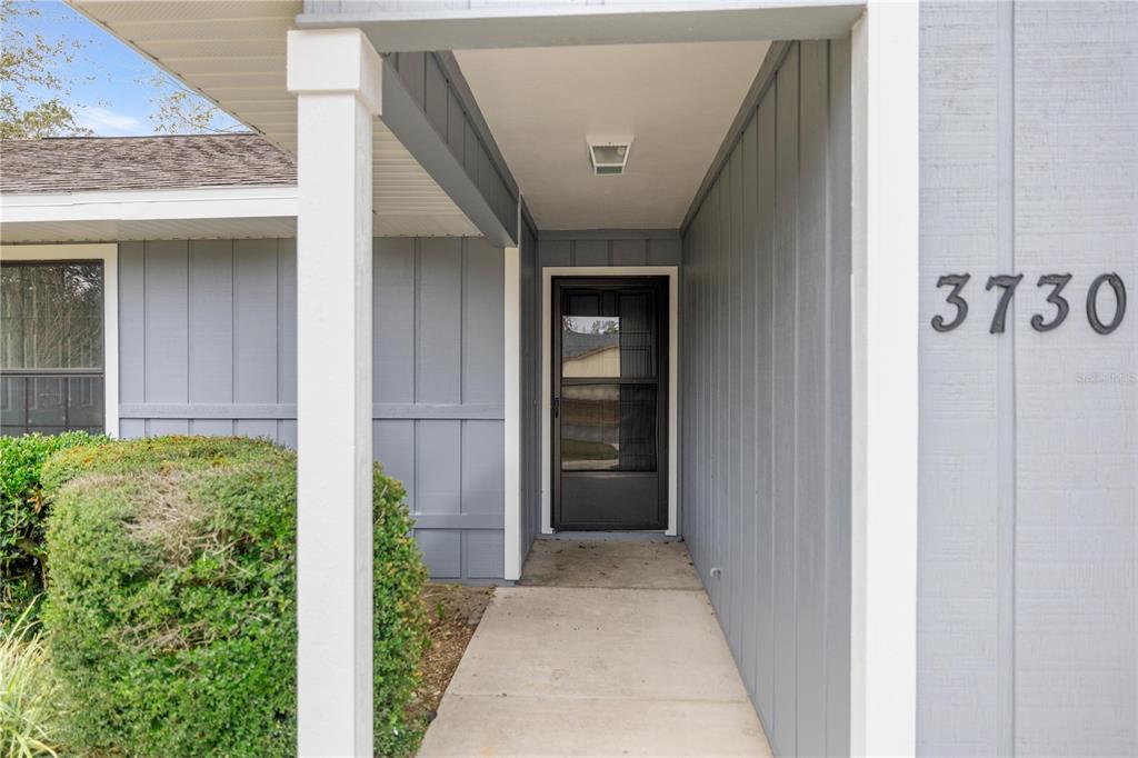 A gray-sided house exterior features a covered entryway with a concrete walkway and storm door — 3730 NW 61ST PLACE, GAINESVILLE, FL