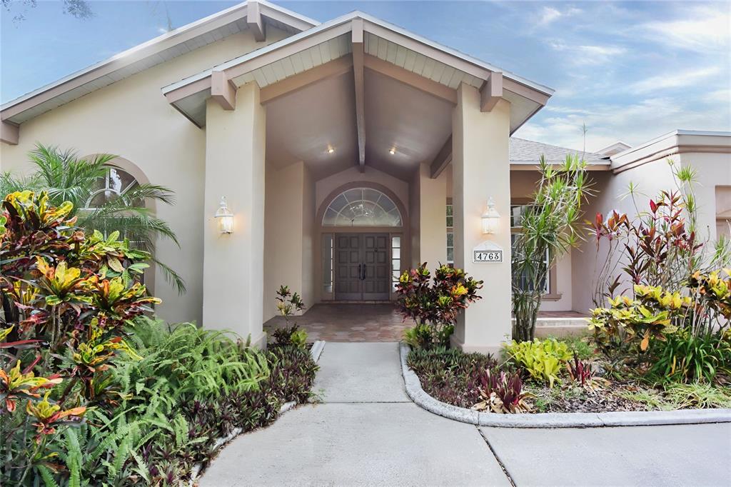 A concrete walkway leads to the covered entryway of a beige house with double doors — 4763 BRAYTON TERRACE N, PALM HARBOR, FL