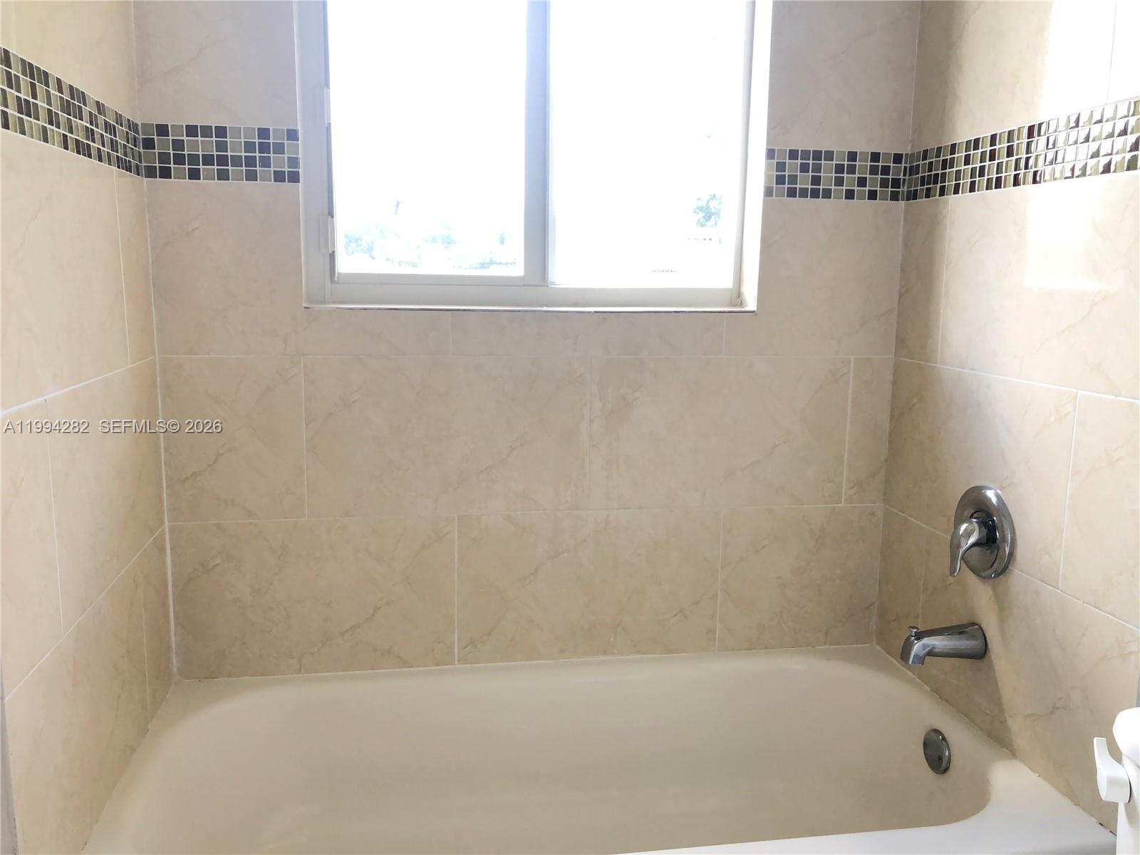 A bathtub with beige tile walls, a window, and a decorative mosaic accent border — 1940 Washington Ave # 11, Opa-Locka FL 33054, OPA-LOCKA, FL