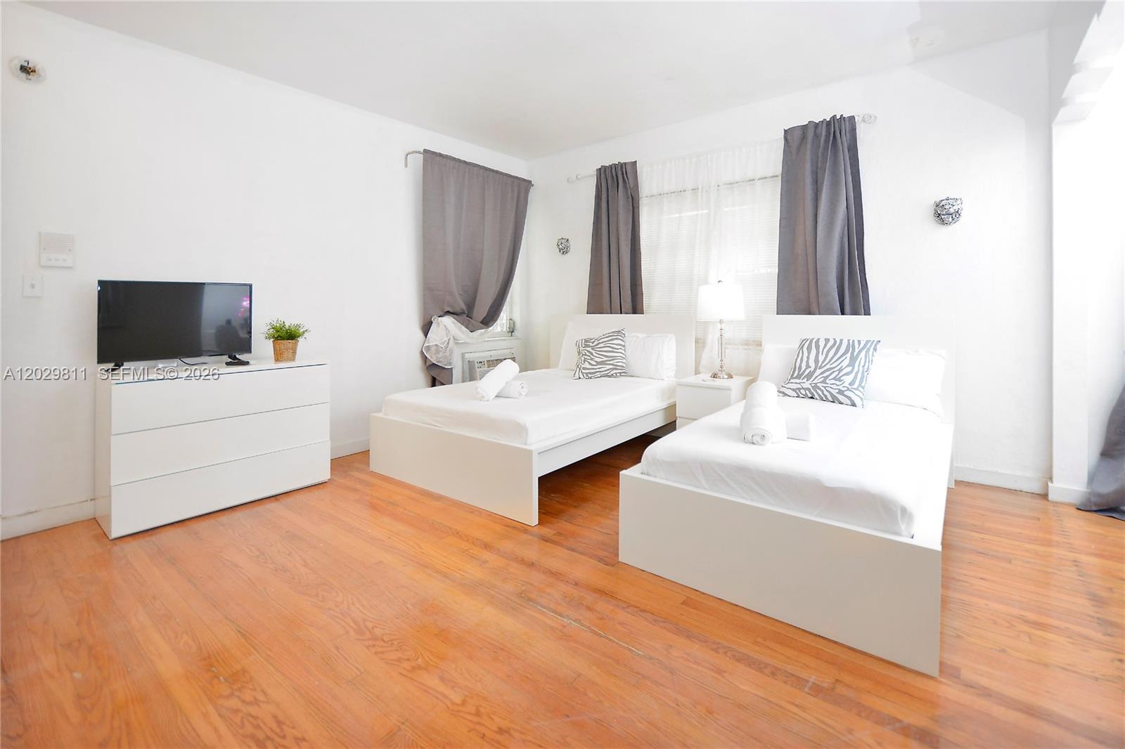 A bedroom features two white beds, a dresser with a television, and hardwood flooring — 1021 Euclid Ave # 1, Miami Beach FL 33139, MIAMI BEACH, FL